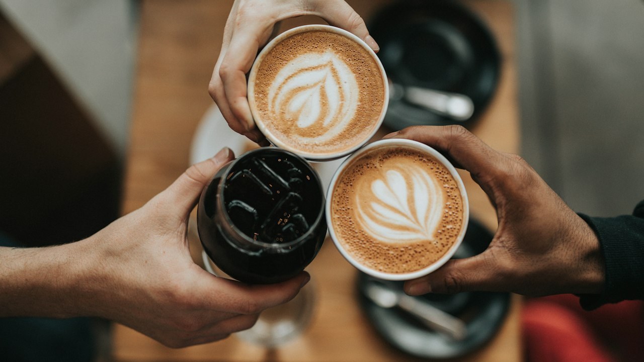 Hands holding two latte cups and an iced coffee over a table